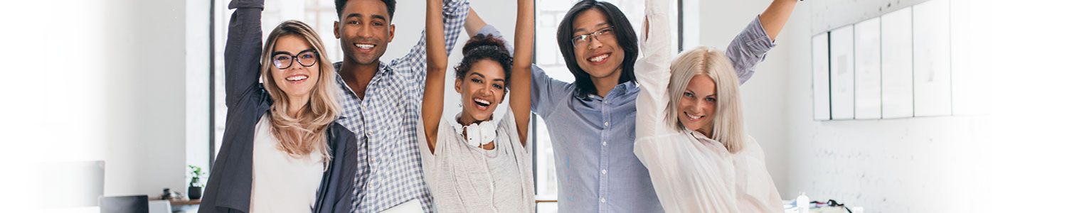 Eine Gruppe fröhlicher und vielfältiger junger Berufstätiger steht mit erhobenen Armen zusammen und feiert in einer Büroumgebung.